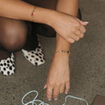 A close-up lifestyle shot of a person's hands and arms wearing delicate gold bracelets and rings, sitting on a textured gray carpet with white-and-black spotted shoes visible in the background.