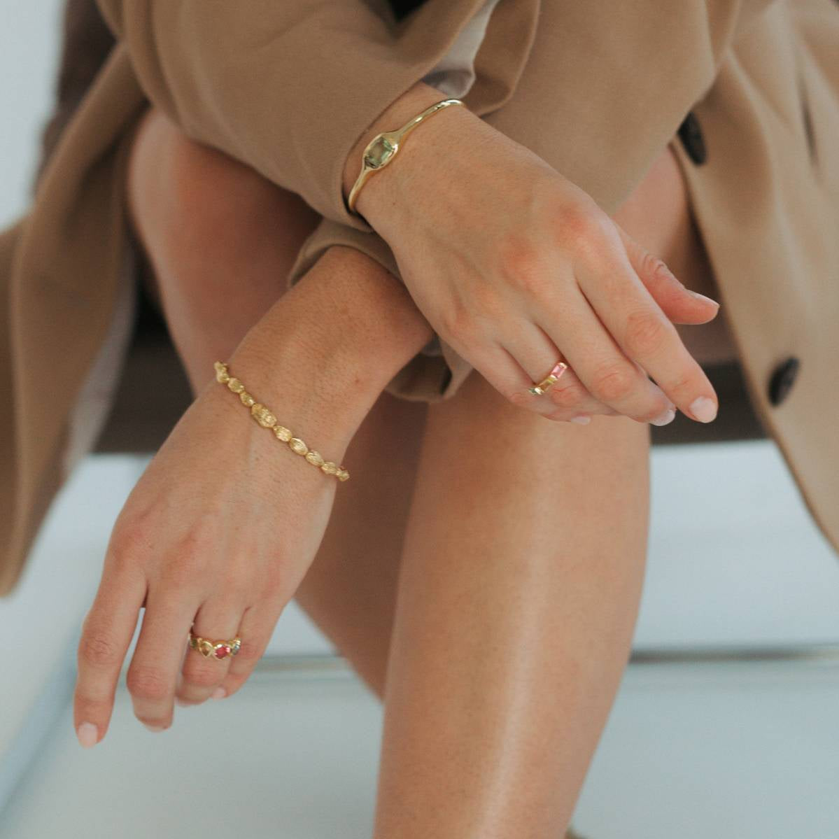A close-up lifestyle shot of a person wearing delicate gold bracelets and rings, dressed in a tan trench coat.