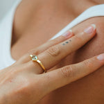Close-up of a hand wearing a gold ring with a green gemstone, touching skin.
