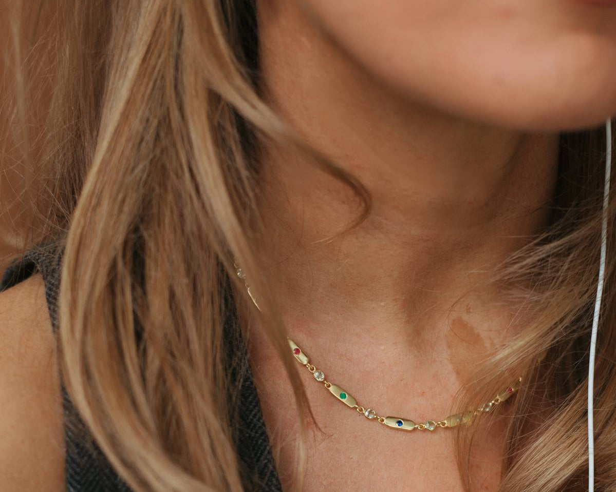 Close-up of a woman wearing a delicate gold chain necklace with small green stone accents, along with a gold hoop earring and white earbud wire.