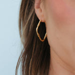 A close-up of a woman's ear wearing a textured, twisted gold hoop earring against a light blue background.