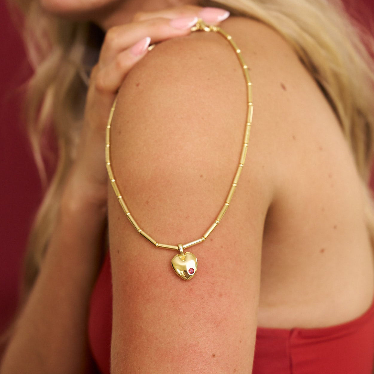 Gold necklace with a heart pendant worn by a person against a red background