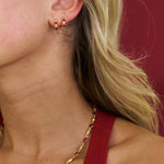 Close-up of a person wearing gold hoop earrings and a gold necklace with red stones against a red background.