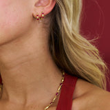 Close-up of a person wearing gold hoop earrings and a gold necklace with red stones against a red background.