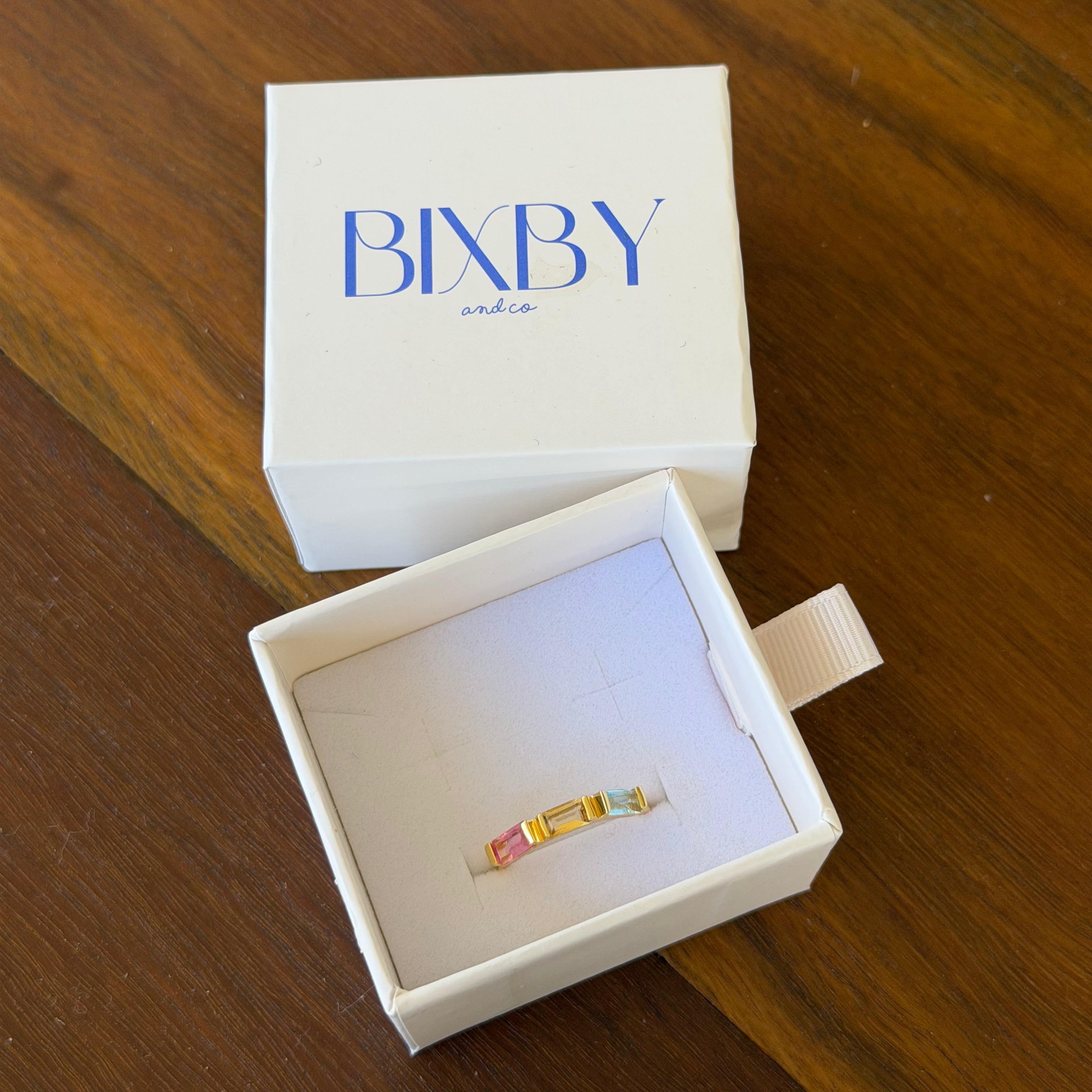 A gold ring with colorful enamel details displayed inside an open white jewelry box, with the white lid featuring the blue 'BIXBY' brand logo placed behind it on a wooden table.