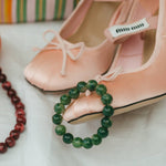 Pink high-heeled shoes with a brand label, green beaded bracelet, and red beaded necklace on a white surface.