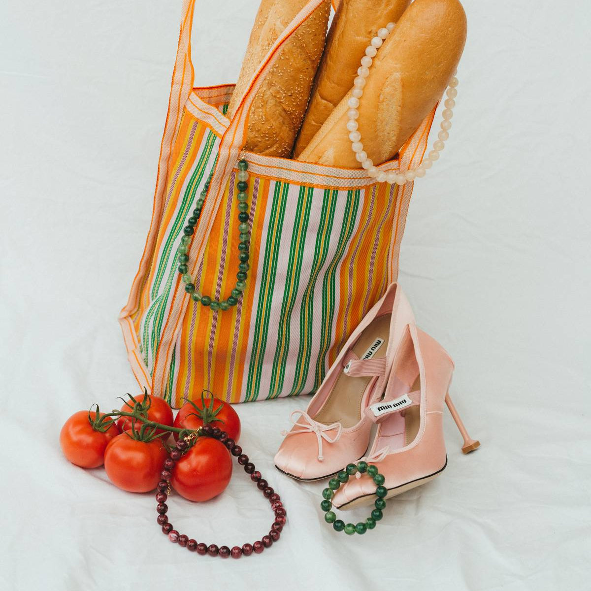 Striped bag with bread, pink high heels, tomatoes, and a necklace on a light background