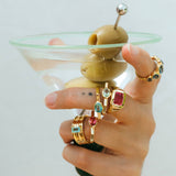 Hand holding a martini glass with a olive and cocktail stick, wearing multiple rings.