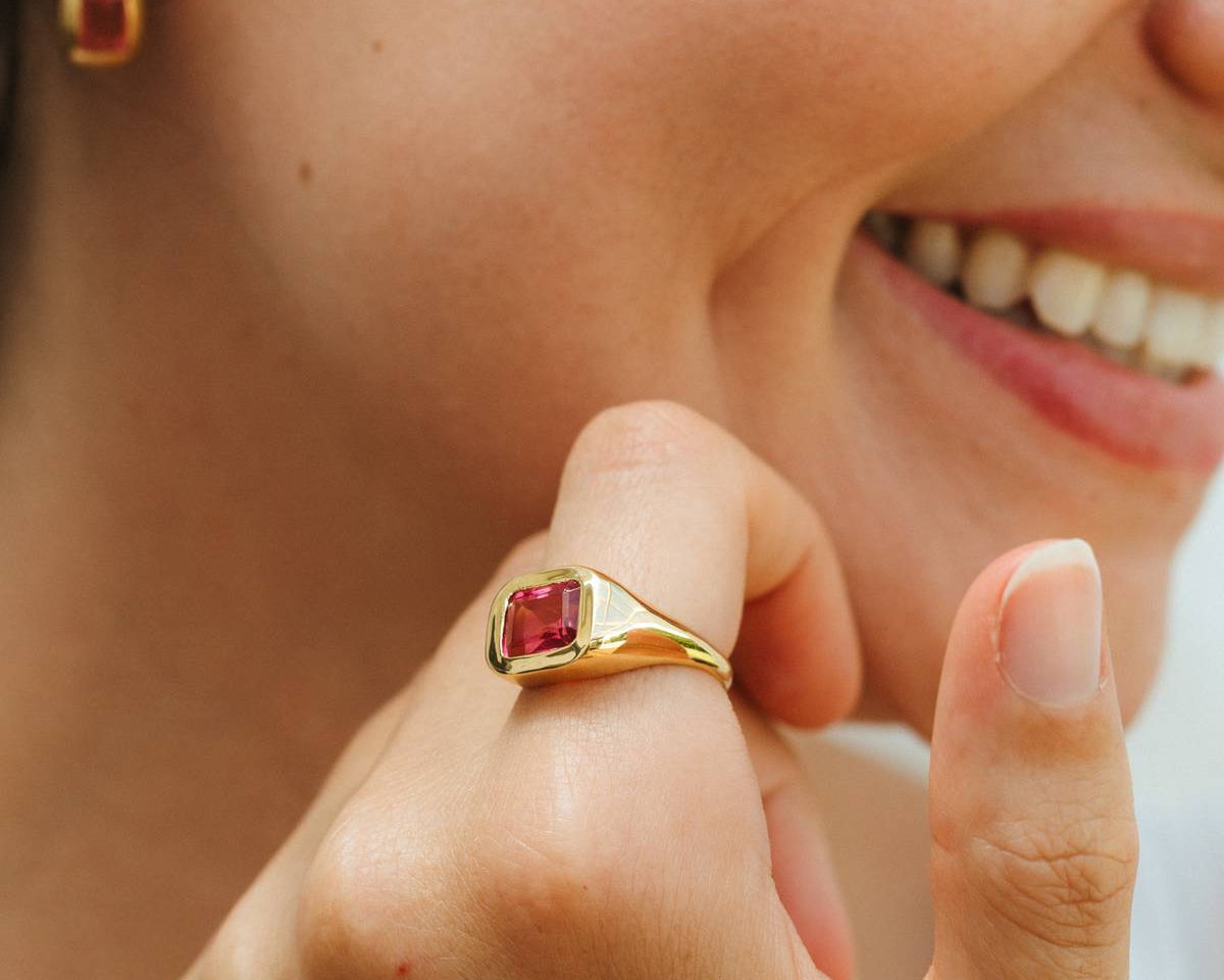Close-up of a woman wearing a gold ring with a pink gemstone, smiling.