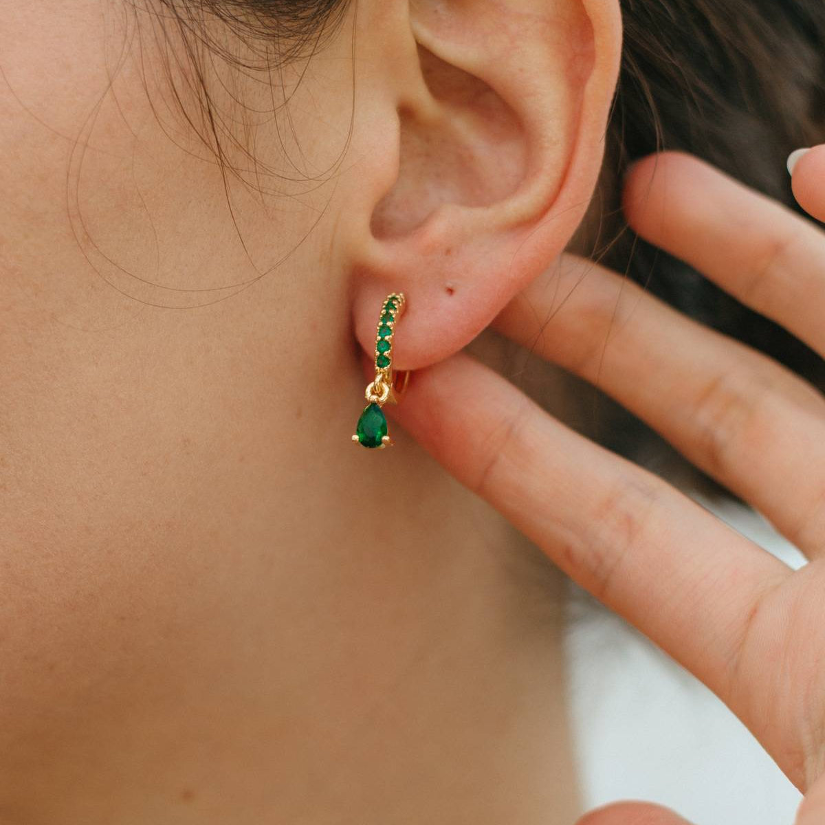 Close-up of an ear wearing a green emerald earring with gold setting, hand touching hair.