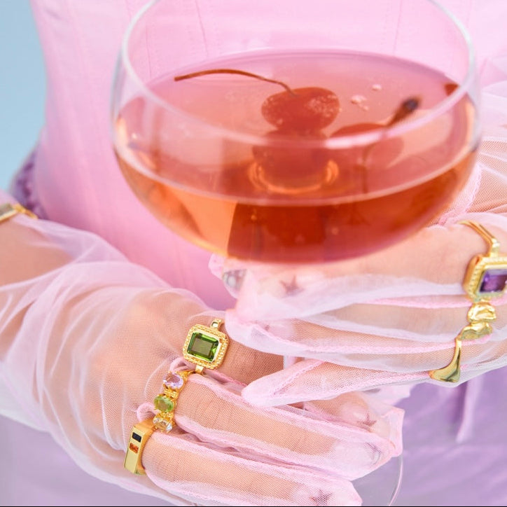 Hand wearing gold rings with gemstones near a glass of pink drink with a cherry on a light purple background